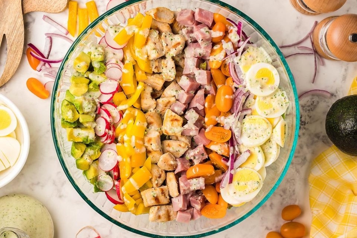 A clear bowl of chef salad with chopped ham, chicken, eggs, tomatoes, radishes, yellow peppers, pickles, red onions, and dressing.