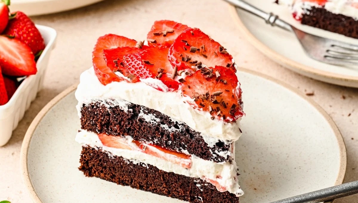 A slice of chocolate cake with white frosting and sliced strawberries on top, on a plate with a fork, surrounded by whole strawberries.