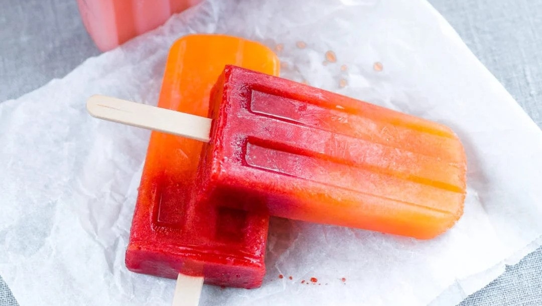 A mold tray filled with freshly made red popsicles and one popsicle lying on a parchment paper beside it.