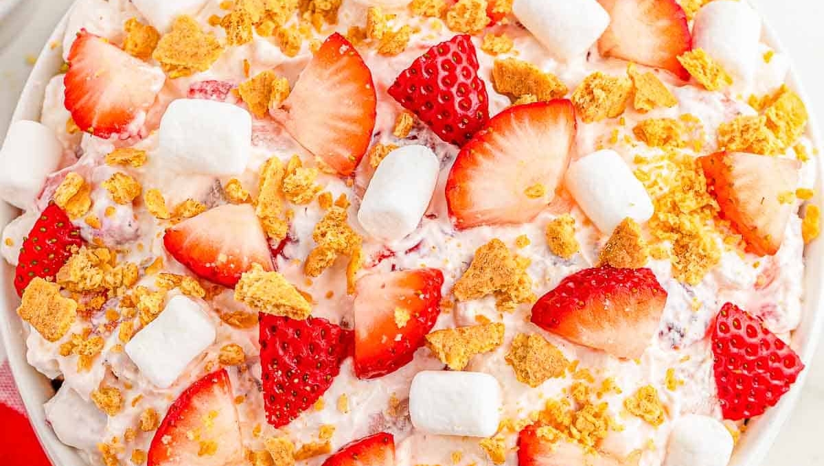 A bowl of strawberry fluff salad topped with fresh strawberry slices, marshmallows, and crushed graham crackers, on a red and white checkered tablecloth.